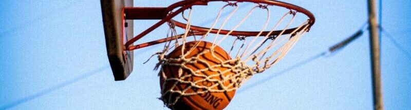 A basketball perfectly swishes through the hoop on an outdoor court against a clear blue sky.